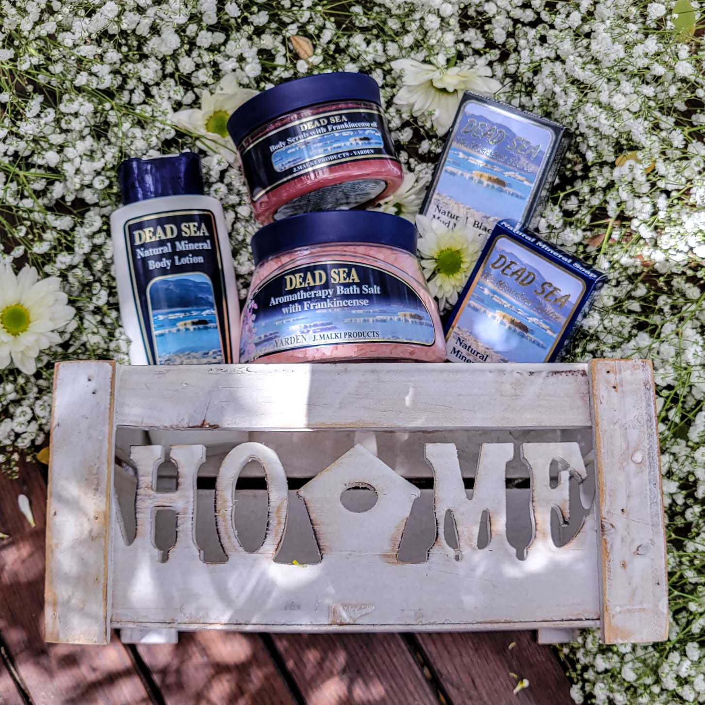 A collection of Dead Sea brand bath and body products, including body cream, facial soap, mineral sponge, body scrub, and a bath salt, arranged on a wooden surface with a decorative piece that reads 'HOME'.