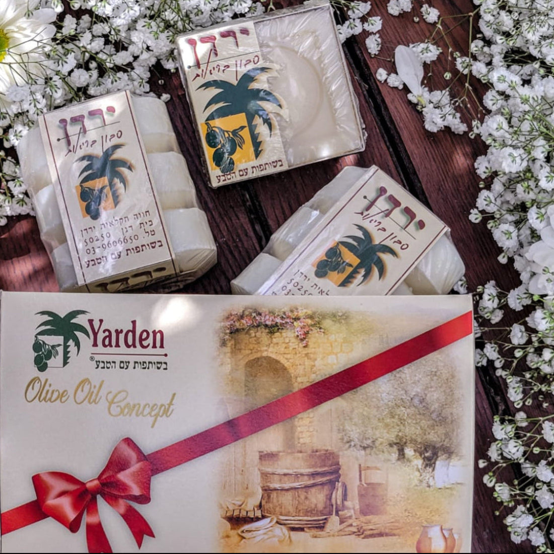 A gift box of Yarden olive oil soap with the packaging displaying an illustration of a palm tree and an olive press, accompanied by two individual soaps wrapped in decorative paper with a red ribbon on top, all presented on a bed of white flower petals.